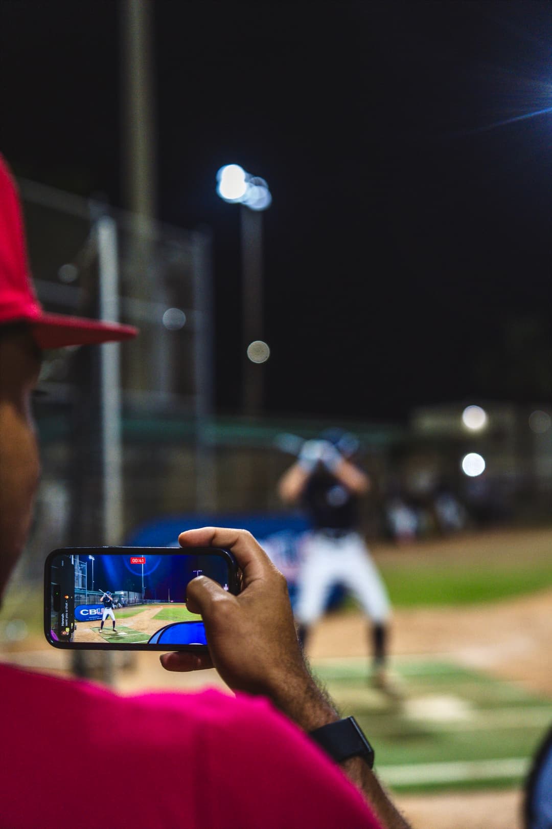 Coach recording batting practice with game action in the background